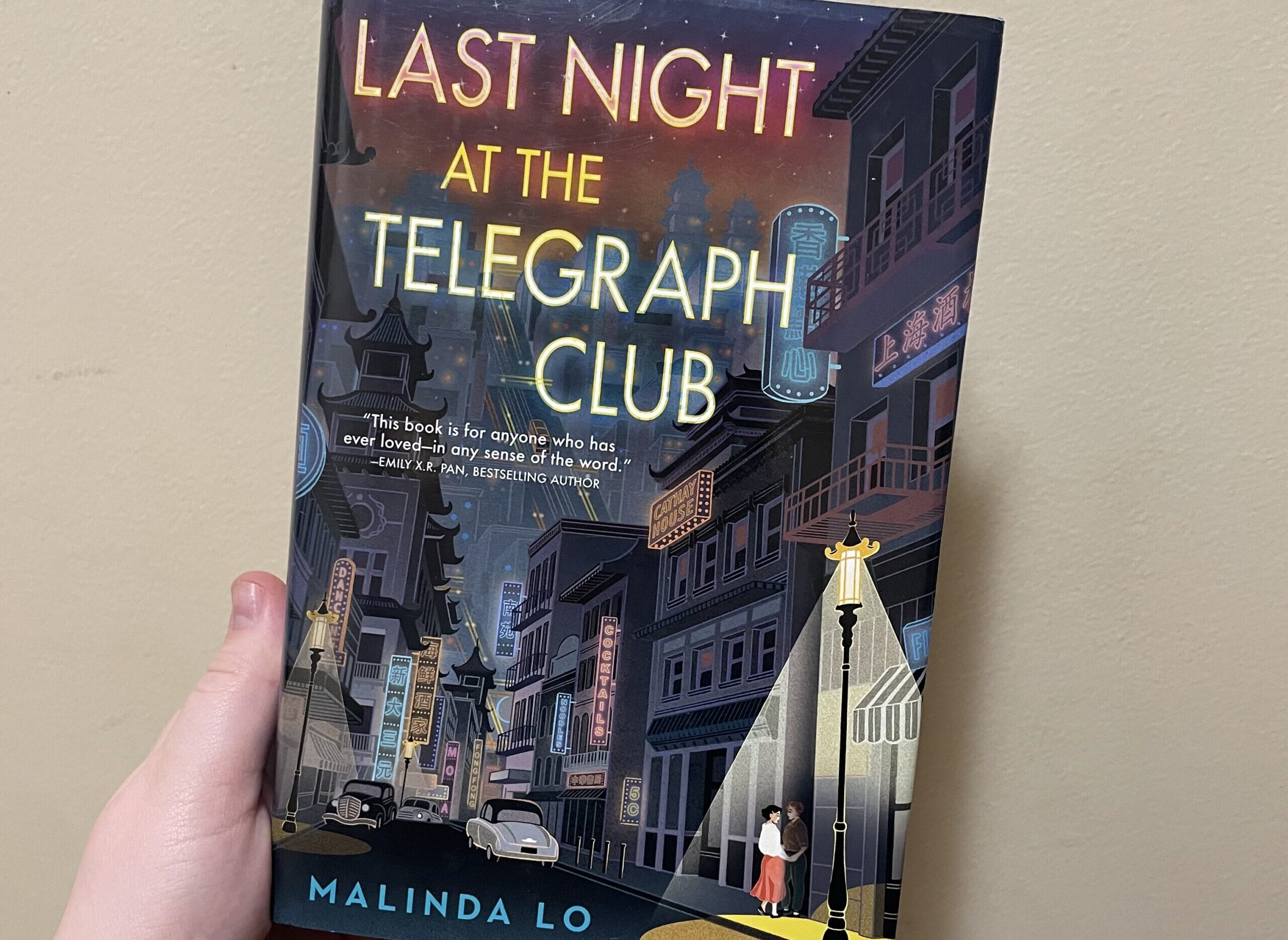 A white hand holds Last Night at the Telegraph Club by Melinda Lo in front of a cream coloured wall. The cover shows a dark street in San Fransisco’s Chinatown. two woman look at each other under a lamppost.